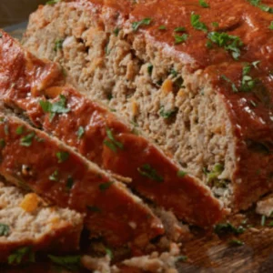 A freshly baked one-pan meatloaf with a glossy, caramelized glaze, sliced and served on a wooden cutting board with mashed potatoes and green beans.

